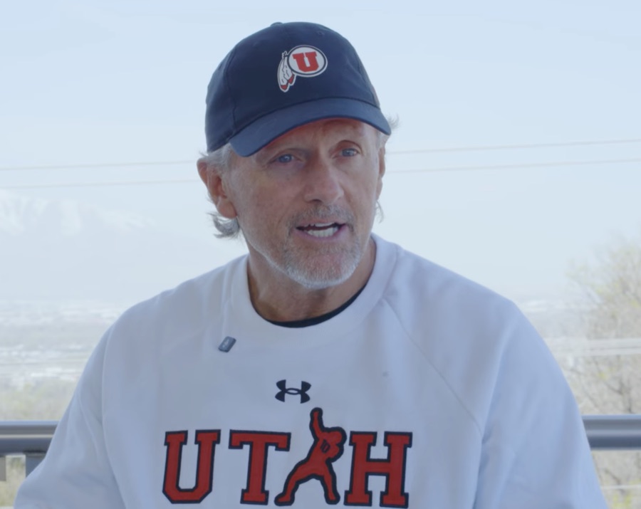 Kyle Whittingham outdoors in a navy Utah cap and a white Utah crewneck sweatshirt, mid-sentence, photographed in May 2025 during his final spring as Utah's head coach, with a hazy valley and distant mountains behind him.