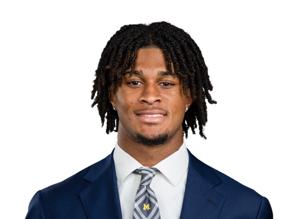 Channing Goodwin in a posed studio portrait against a white background, wearing a navy suit jacket over a white shirt and a gray tie striped in maize and navy, with a small maize block M low on the tie. No jersey and no number are in the frame.