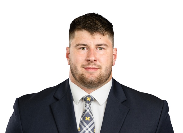 Manuel Beigel, Michigan offensive lineman, in a posed roster portrait against a white background, wearing a navy suit jacket, white shirt and a navy and maize plaid tie carrying a small block M.
