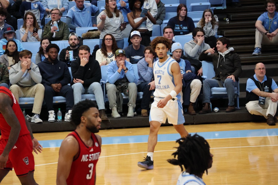A guard in a North Carolina home white uniform, jersey reading NORTH CAROLINA over the number 7, standing in the frontcourt during a game against NC State, with two red-jerseyed NC State players in the foreground and a packed courtside crowd in Carolina blue behind him.