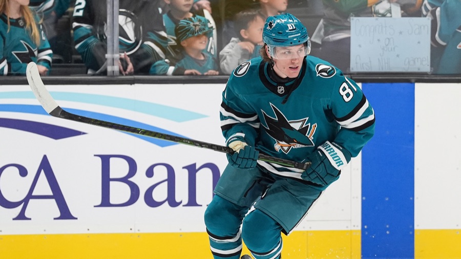 Adam Gaudette skating up ice in San Jose's teal home sweater, number 81 visible on his right sleeve and on the back of his helmet, stick in both hands and head up. A Sharks crowd sits behind the glass.