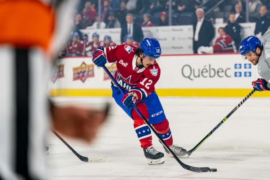 Alex Barre-Boulet carrying the puck in a red and blue AHL All-Star sweater with a white 12 on the chest, an opponent in grey closing from the right. The 2023 AHL All-Star Classic at Laval, February 6, 2023, when he was a Syracuse Crunch player. It is not a San Jose Sharks or San Jose Barracuda uniform, and no photograph of him in either exists: he signed with San Jose on July 1, 2026 and has not yet played a game for the club.