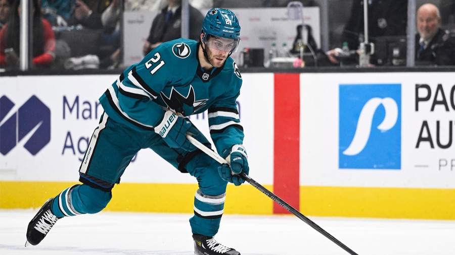 Alexander Wennberg in San Jose's teal home sweater, leaning into a stride with the puck ahead of his stick blade, the boards and a rink-side advertising panel behind him.