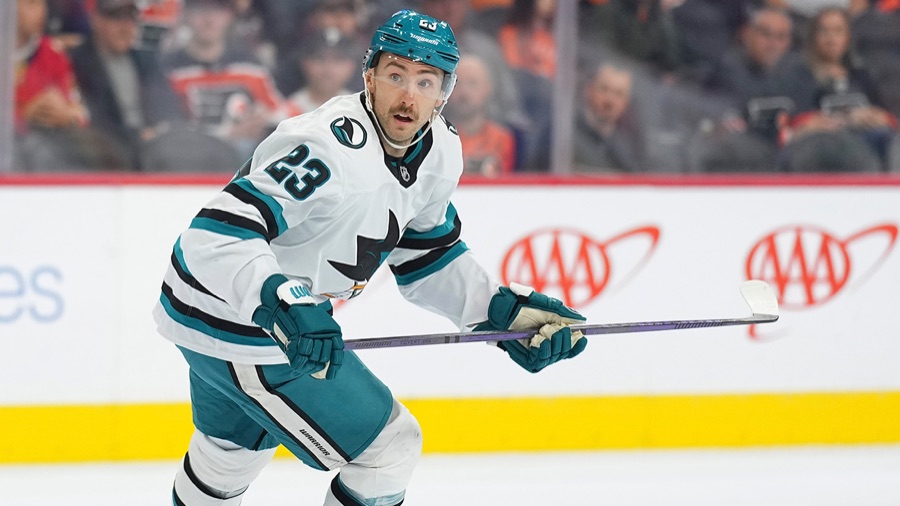 Barclay Goodrow in San Jose's white road sweater, the teal and black Sharks fin crest at the chest and the number 23 on his shoulder, skating up ice with his stick out in front of him and the boards behind him.