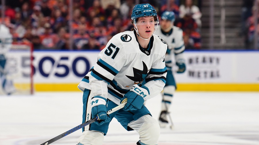 Collin Graf in the Sharks' white road sweater, knees bent and stick on the ice, tracking the play in Edmonton with a teammate behind him along the boards.