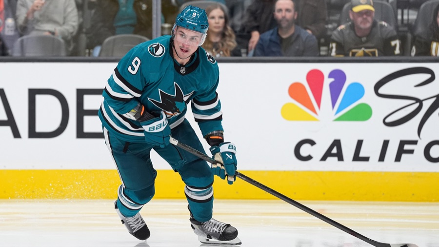 Dmitry Orlov in the Sharks' teal home sweater, number 9, carrying the puck up the ice and looking to his right, with the boards and a crowd behind him