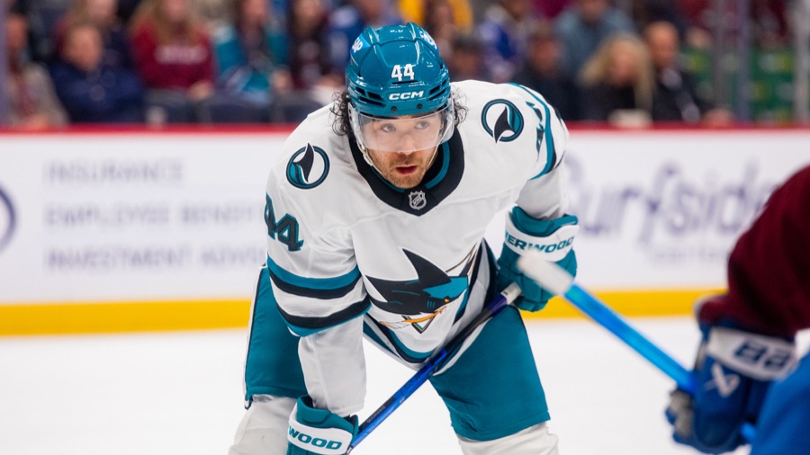 Kiefer Sherwood in a white San Jose Sharks road sweater with teal trim, crouched over his stick waiting for a faceoff, an opponent in dark colours blurred at the right edge of the frame.