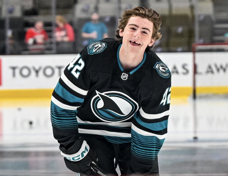 Luca Cagnoni in a black San Jose Sharks sweater, number 42 visible on the shoulder and the sleeve, crouched on the ice in warmups and laughing, with the boards and a sparse lower bowl behind him. The photograph carries no date from its source.