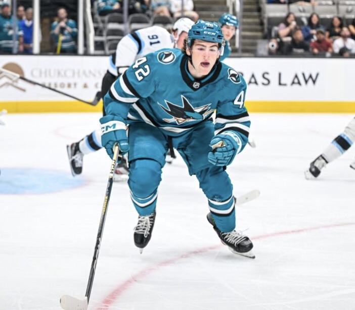 Luca Cagnoni in San Jose's teal home sweater with the Sharks fin crest, skating hard through the neutral zone with the puck ahead of his stick, an opponent in white behind him and a full arena crowd beyond the boards.