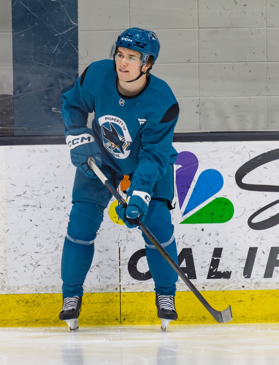 Macklin Celebrini at a San Jose Sharks practice in April 2026, in the teal practice sweater with the Property of San Jose fin crest at the chest and a teal Sharks helmet, standing on the ice with his stick in front of him and the boards behind him.