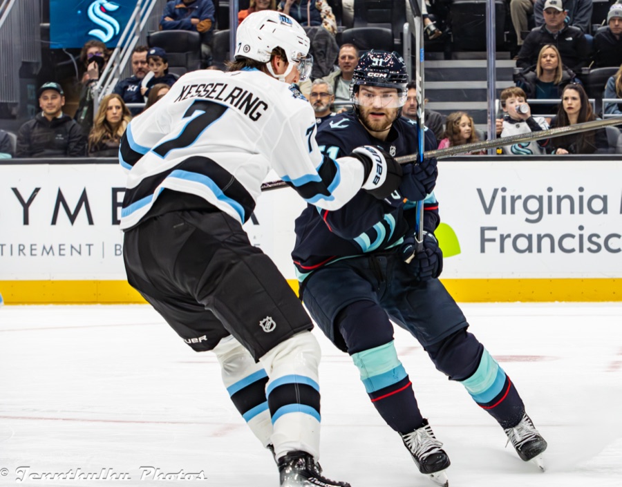 Michael Kesselring in a white Utah road sweater with his surname across the shoulders, seen from behind as he closes on a Seattle forward carrying the puck up the boards.