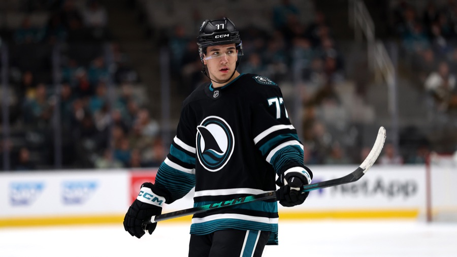 Michael Misa on the ice in a black Sharks sweater carrying the wave crest, with teal and white striping on the sleeves, holding his stick across his body and looking off toward the right of the frame. A white 77 is legible on the front of his black helmet and again on his right shoulder. An out-of-focus arena crowd and a rink-board advertisement fill the background.