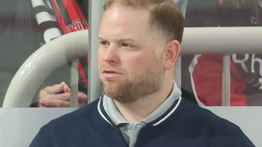 Ryan Warsofsky behind a bench in a navy USA Hockey quarter-zip over a grey collar, looking to his right past the boards. Rink glass and a white railing behind him. A May 2025 photograph from Germany against the United States, where he was head coach of the American team, sixteen months before the extension described here.