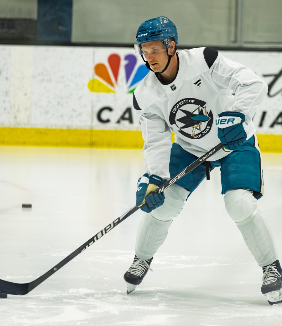 Ty Dellandrea in a white San Jose practice sweater with the Property of San Jose crest, teal helmet and teal gloves, leaning into a stride and reaching for a loose puck at practice