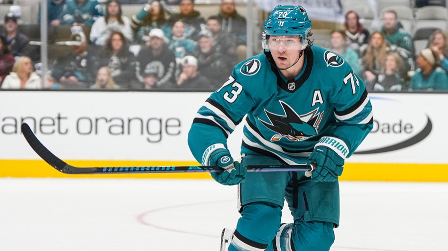 Tyler Toffoli in a San Jose Sharks teal home sweater with an alternate captain's A on the chest, upright and gliding with both hands on his stick, a full crowd behind the boards.