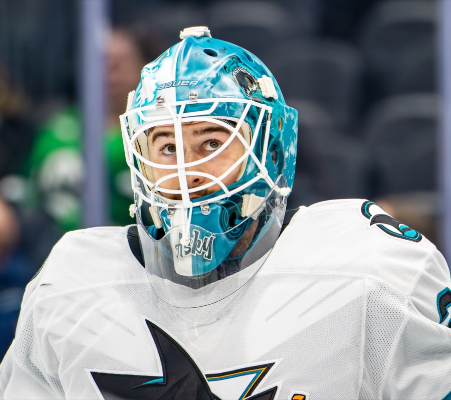 A close portrait of Yaroslav Askarov in a white Sharks sweater with the teal, black and white shark crest on the chest, his head tilted up and turned away from the camera. He wears a teal goaltender mask with a white cage, and the word ASKY is painted in white script across the chin. Out-of-focus arena seating fills the background. The photograph is filed under the 2024-25 San Jose season.