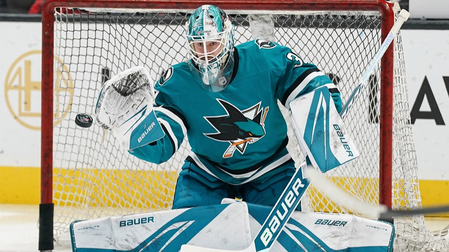 Yaroslav Askarov in the Sharks' teal sweater, upright in the crease with the puck at his glove hand and the net and the arena boards behind him.