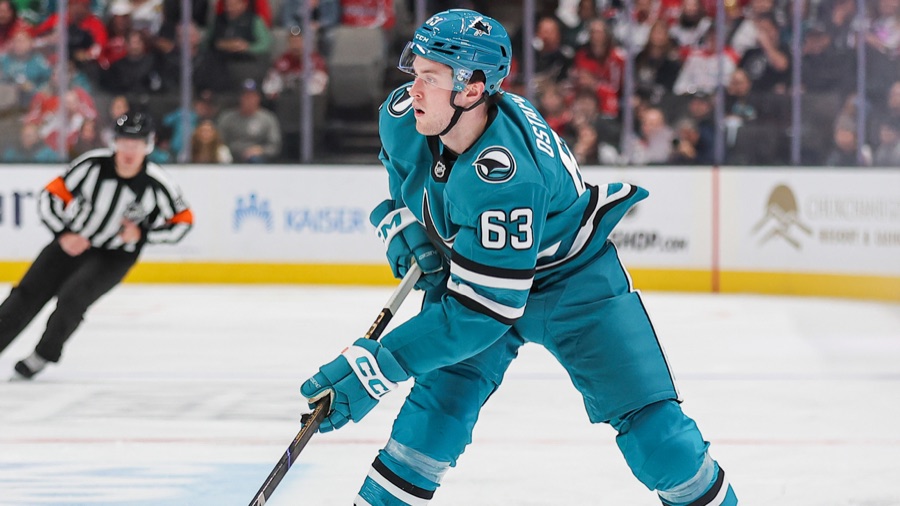 Zack Ostapchuk in San Jose's teal home sweater, the Sharks fin crest at the chest and his surname across the shoulders, leaning into a stride with his stick down and the boards behind him.