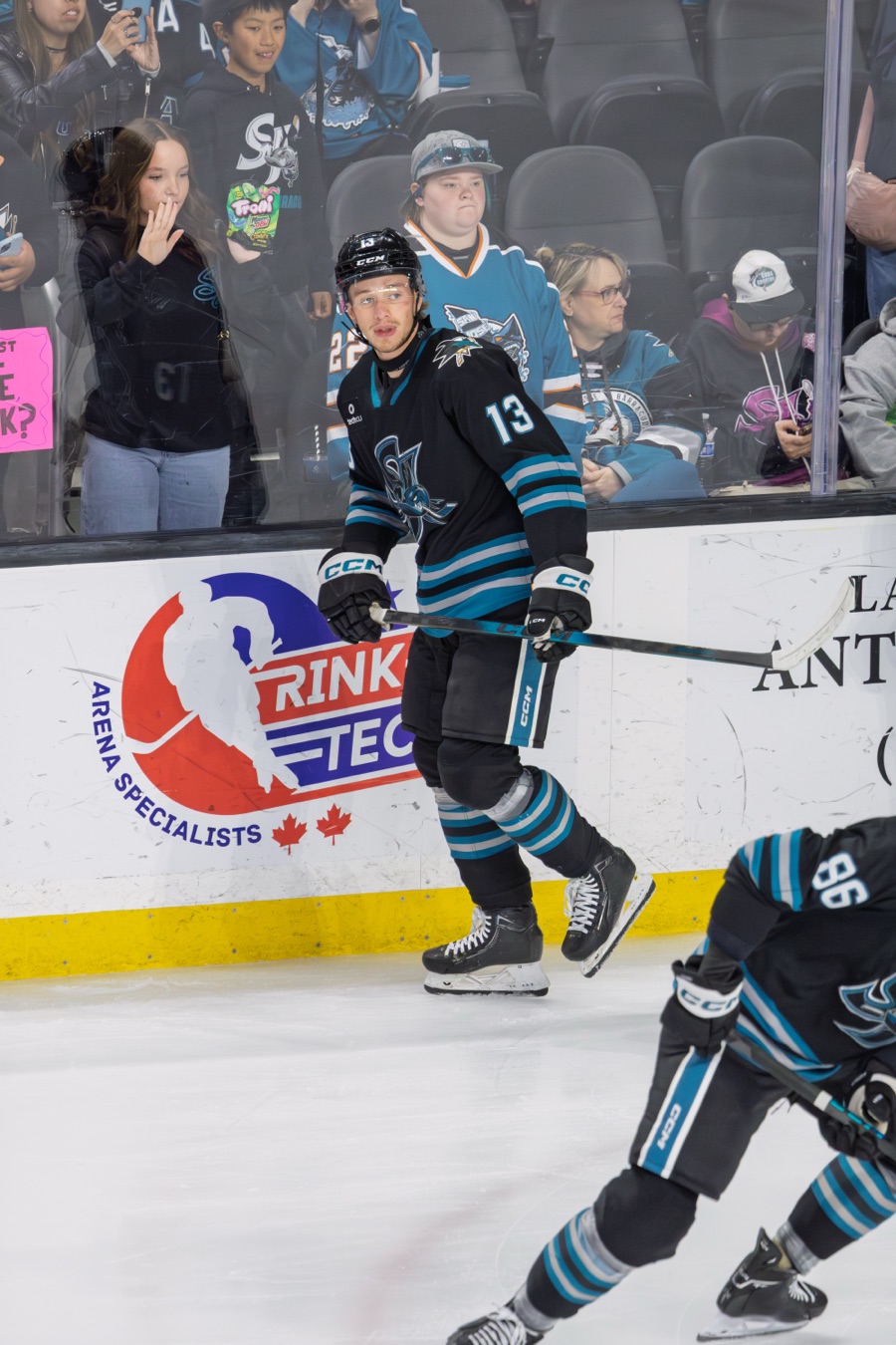 Quentin Musty gliding along the boards in the San Jose Barracuda's black home sweater with teal and grey striping, stick held in both hands, spectators visible behind the glass.