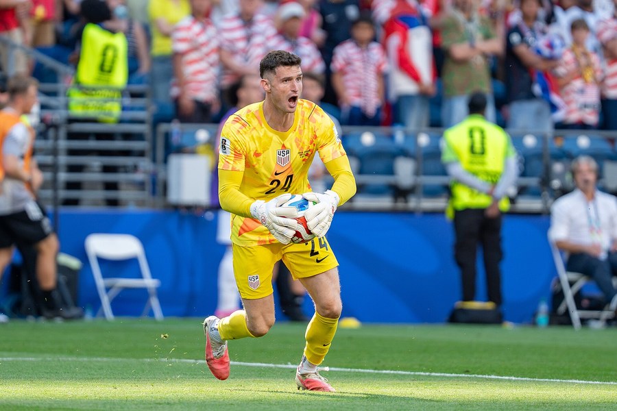 Matt Freese, in the yellow United States goalkeeper kit, runs with the ball held in both gloves against Belgium, with stewards and spectators behind him.
