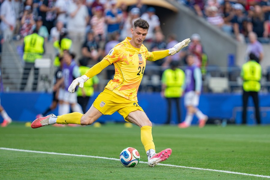 Matt Freese, in a yellow United States goalkeeper kit, strikes a ball along the ground with his right foot, arms spread, during the World Cup match against Belgium in July 2026, with stewards in yellow vests and spectators behind him.