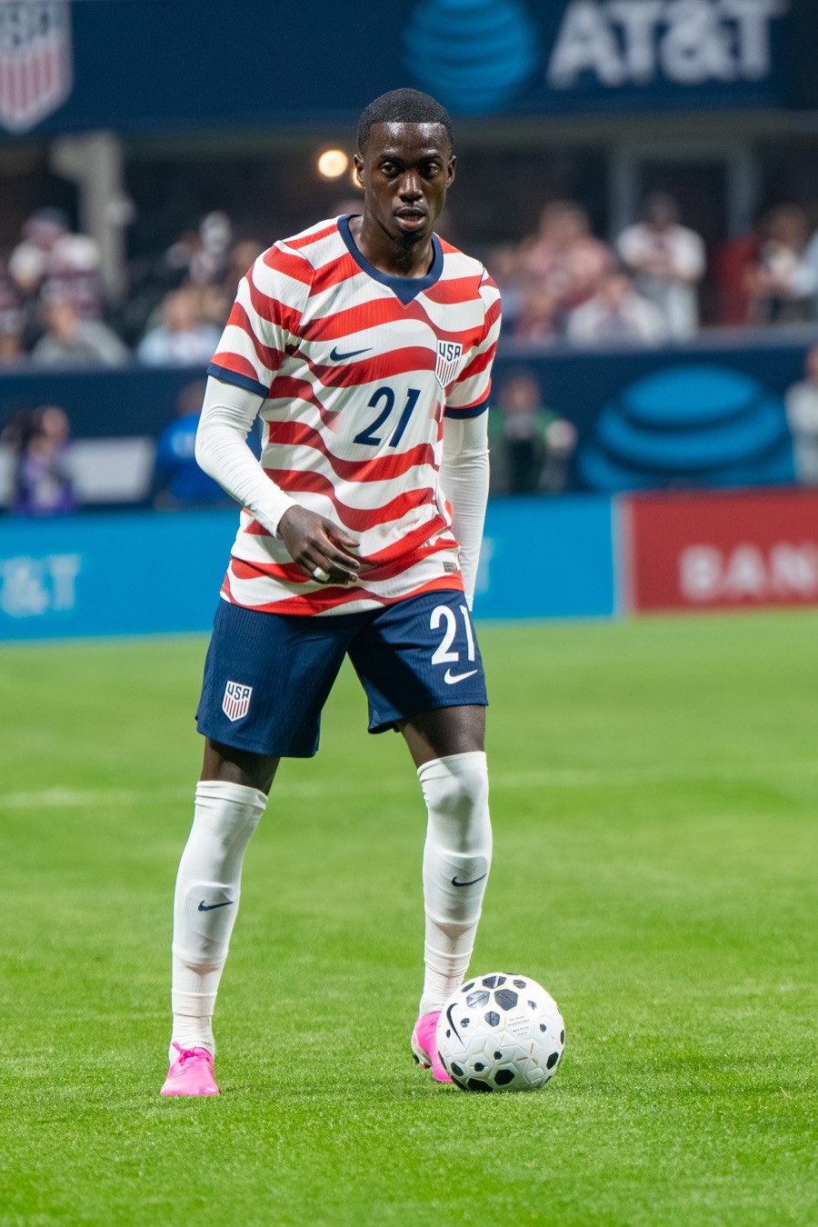 Timothy Weah in the red-and-white striped United States shirt, standing over the ball on the grass in a covered stadium, playing against Belgium in Atlanta in March 2026.