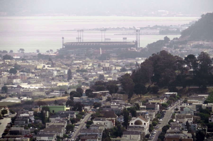 Candlestick Park photographed from a hillside to the west in 2014, the year before its demolition was completed. The ballpark sits low and pale in the middle distance above the rooftops of the Bayview, its light towers along the rim, with San Francisco Bay and a bank of haze behind it.
