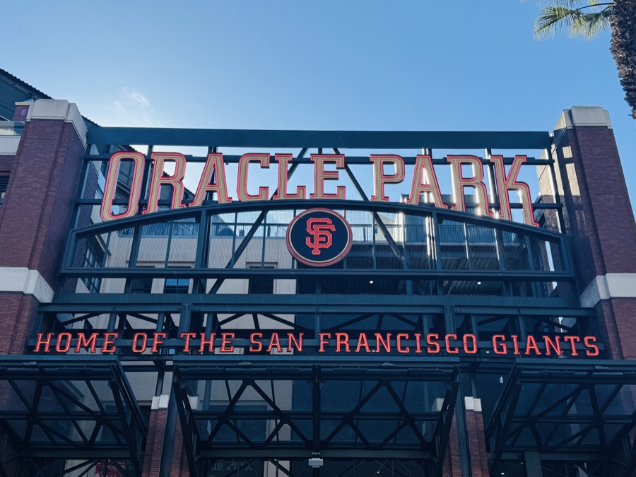 The gate sign above the entrance to Oracle Park in San Francisco, reading ORACLE PARK over the interlocking SF logo, with HOME OF THE SAN FRANCISCO GIANTS in orange lettering below it against a clear blue sky.