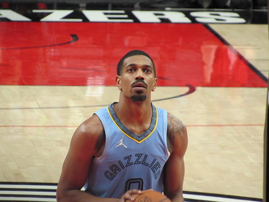 De'Anthony Melton at the free-throw line in a pale blue Memphis Grizzlies jersey, looking up at the rim, with the Portland court markings behind him. The photograph is from October 2021, four seasons before he signed his current Golden State contract.