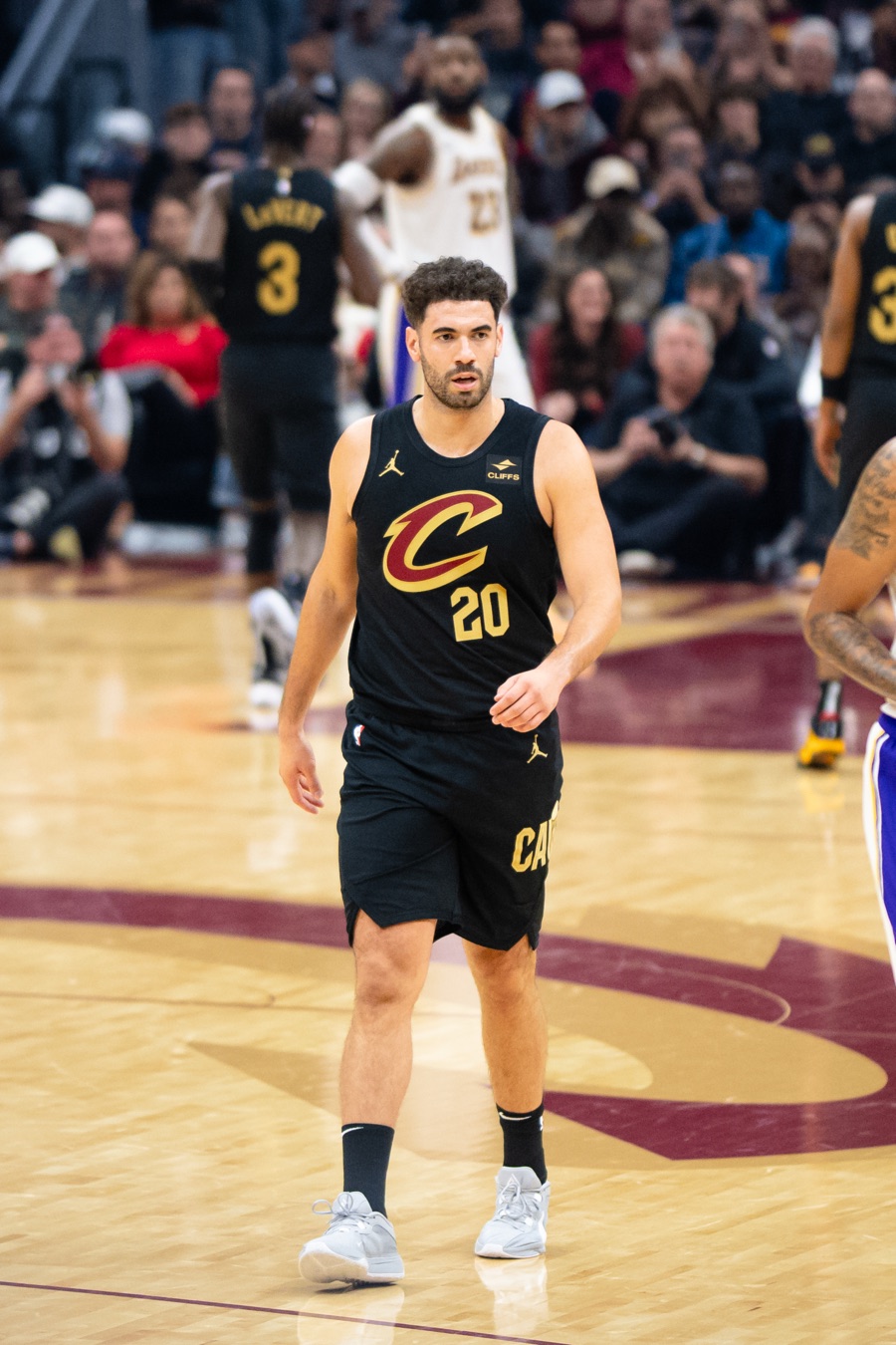 Georges Niang jogging up the floor in Cleveland's black alternate jersey, the gold C and 20 on his chest and a CLIFFS patch at the shoulder, with LeBron James in Lakers white behind his right shoulder. This is a photograph from November 2023, when Niang played for Cleveland.