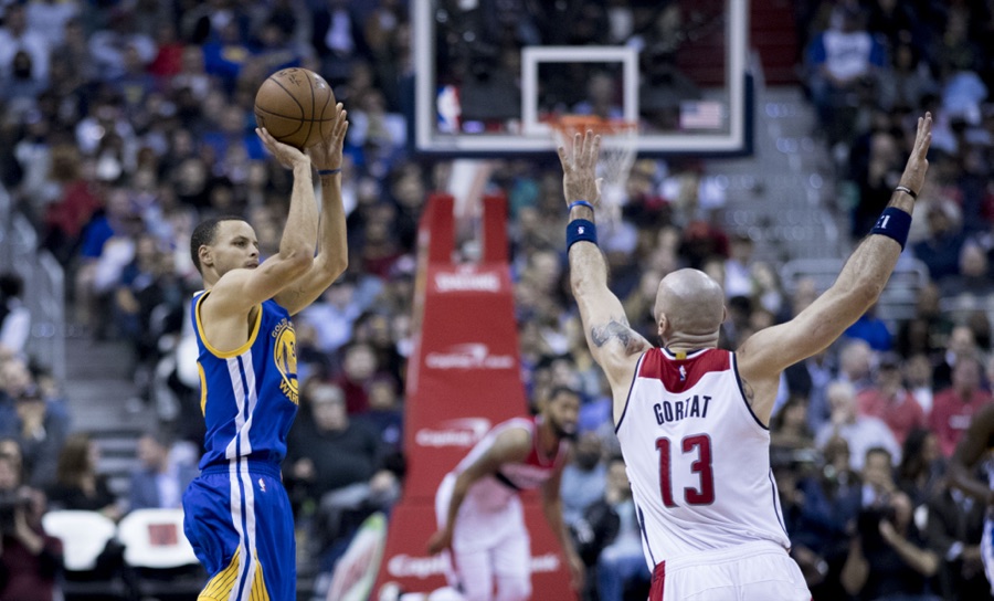 Stephen Curry, in the Golden State road blue jersey, releasing a jump shot from distance as Washington's Marcin Gortat closes with both arms raised. Photographed in February 2017.