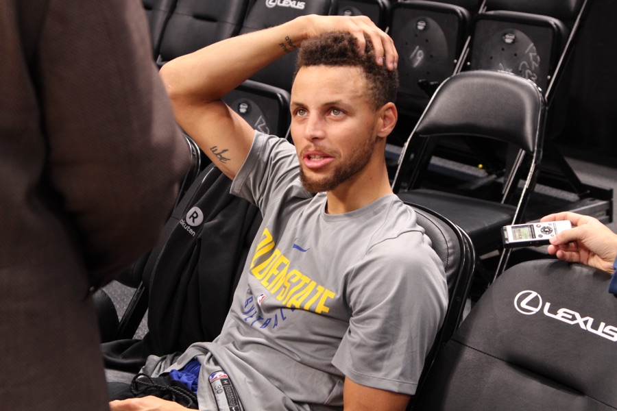 Stephen Curry in a gray Golden State Warriors shooting shirt sits courtside answering a question while a reporter's handheld recorder is held into the frame