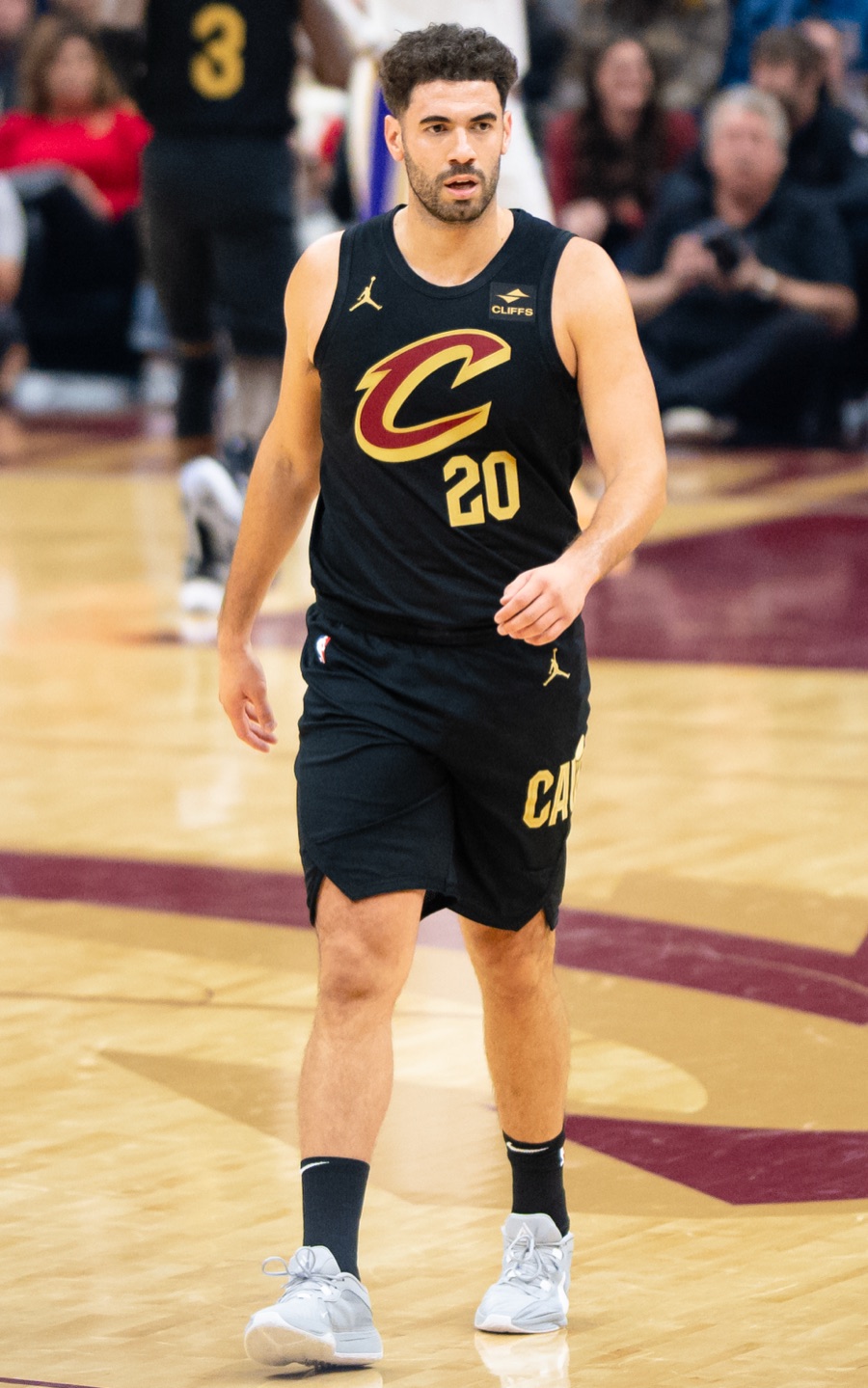 Georges Niang walking up the floor in the Cleveland Cavaliers' black alternate jersey, gold wordmark across the chest and a CLIFFS patch at the shoulder, on the maroon and gold floor of the club's arena with spectators behind him.